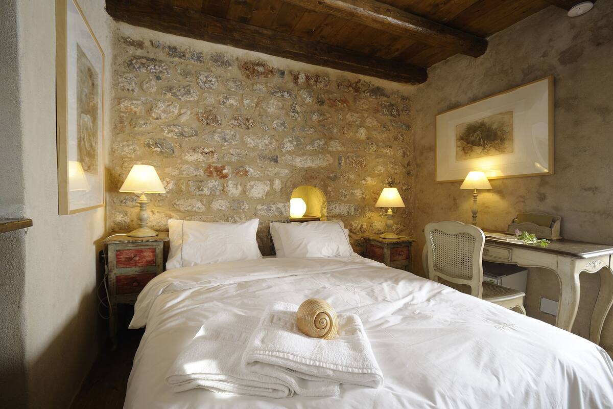 A bedroom with a stone wall, a bed with white linens, two bedside lamps, a wooden desk, and a chair.