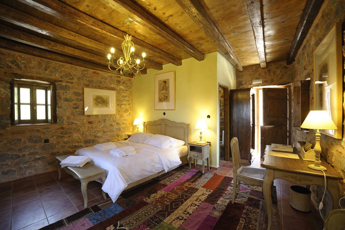 A rustic bedroom with a stone wall, wooden ceiling beams, a bed with white linens, a chandelier, and a desk with a lamp.