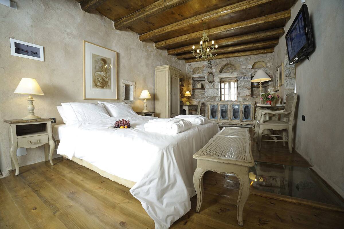 A bedroom with a bed, bedside tables, a chandelier, a television, and a stone wall.