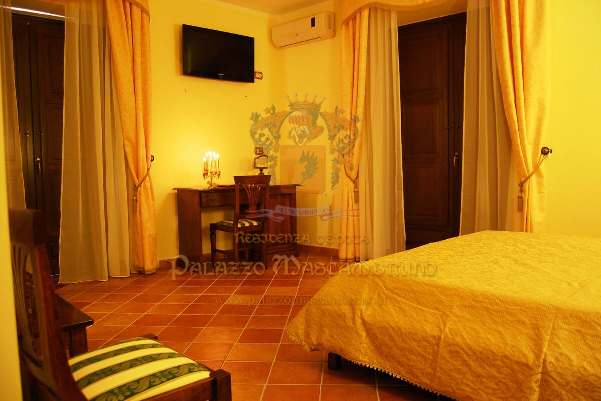 Quarto de hotel com cama, escrivaninha com luminária, uma cadeira e cortinas.