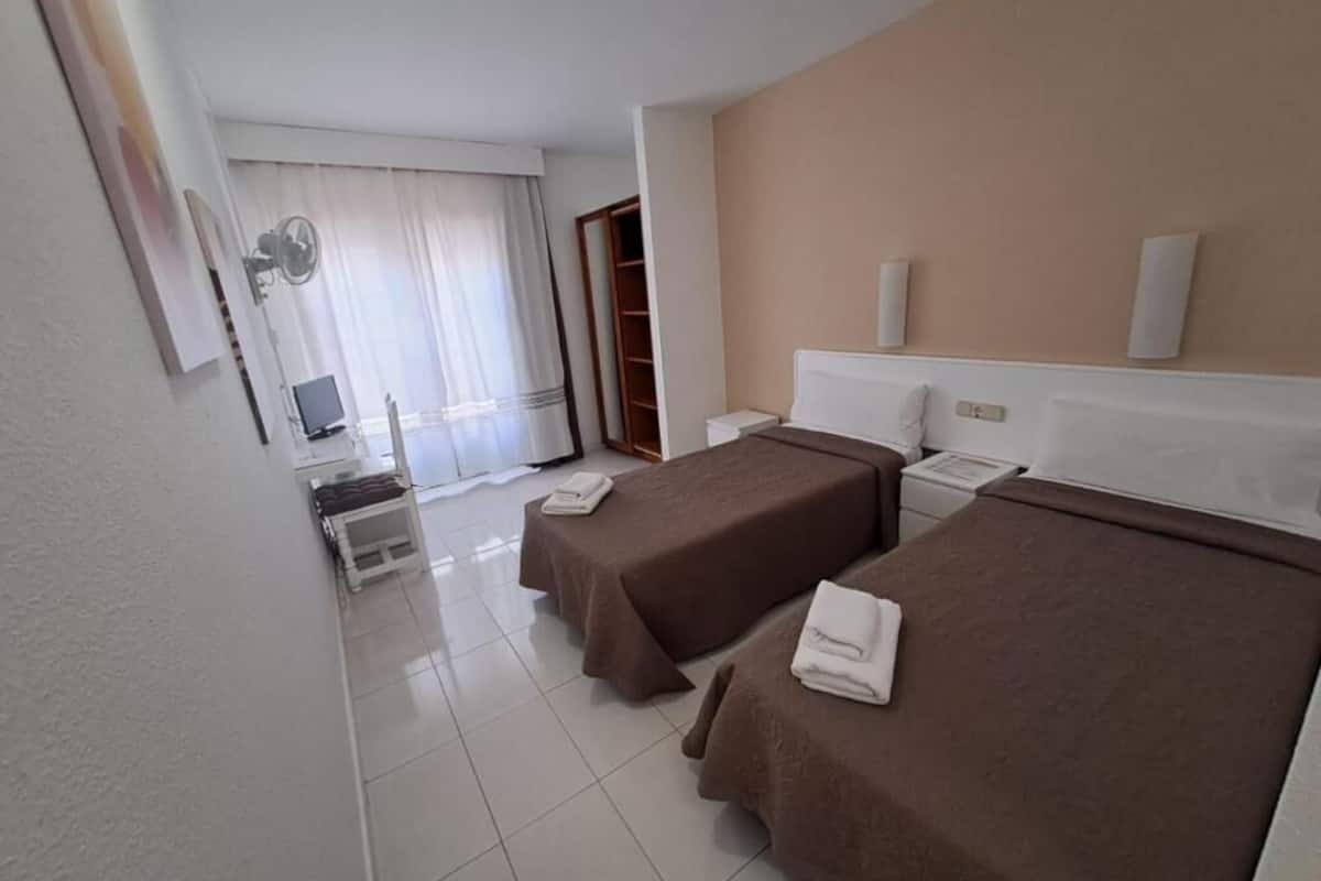 Habitación de hotel con dos camas, un escritorio con ordenador, un ventilador y una ventana con cortinas.