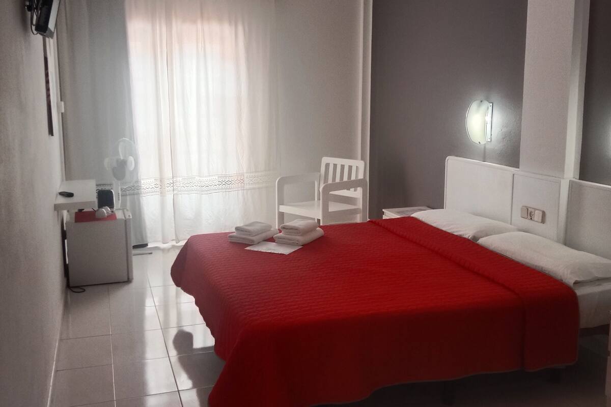Una habitación de hotel moderna con una cama grande, una mesita de noche, una silla y un ventanal con cortinas.