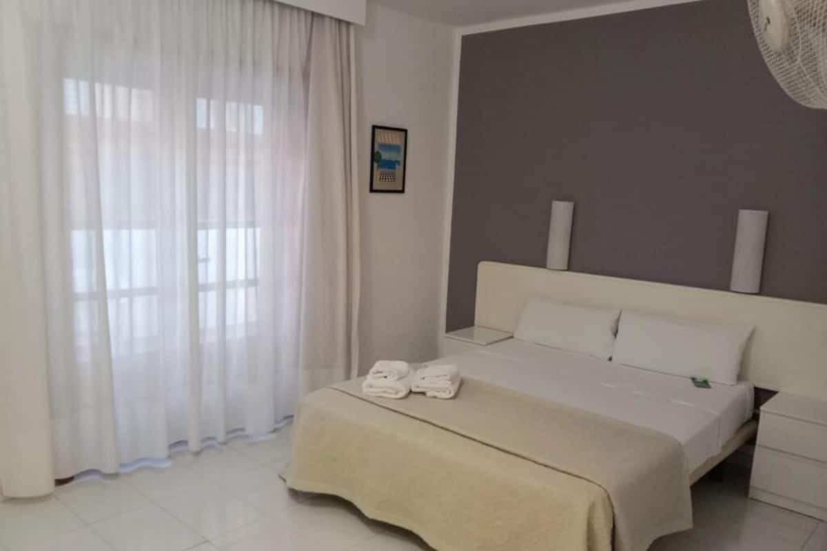 Habitación de hotel con cama, mesita de noche, cortinas translúcidas y ventilador de pared.