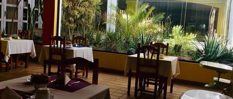 Un restaurante con mesas y sillas, rodeado de plantas y vistas a edificios.