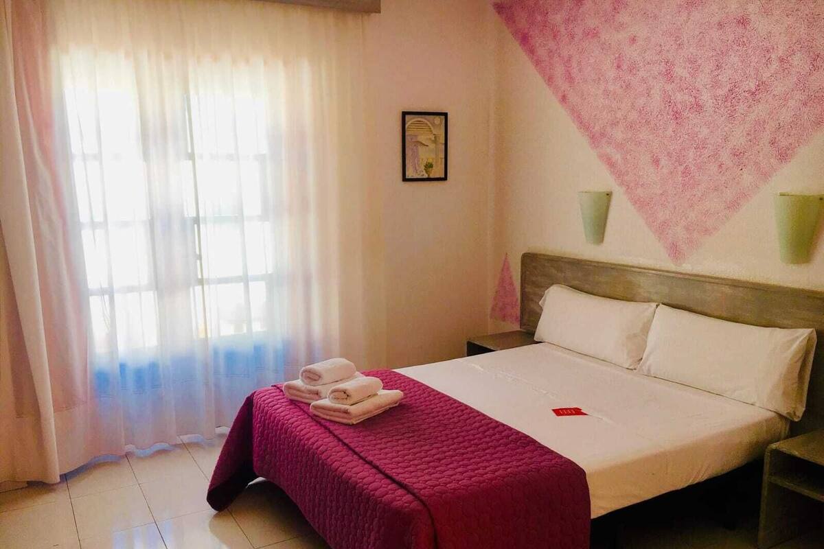 Una habitación de hotel con cama, mesita de noche, ventana con cortinas y un adorno en la pared.