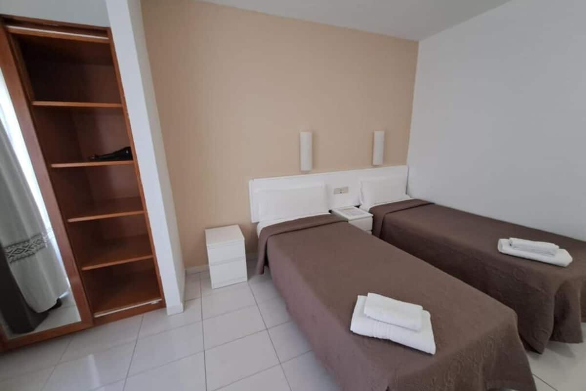 Una habitación de hotel con dos camas individuales, un armario de madera, una mesita de noche blanca y una pared color beige.
