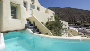 Un área junto a la piscina con una piscina azul cristalina, una escalera que conduce a un edificio de fachada blanca, y montañas al fondo.