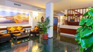 A lobby with a reception desk, seating area with sofas and a coffee table, a large painting on the wall, and a potted plant. - Hotel Galera (Sant Antoni de Portmany)