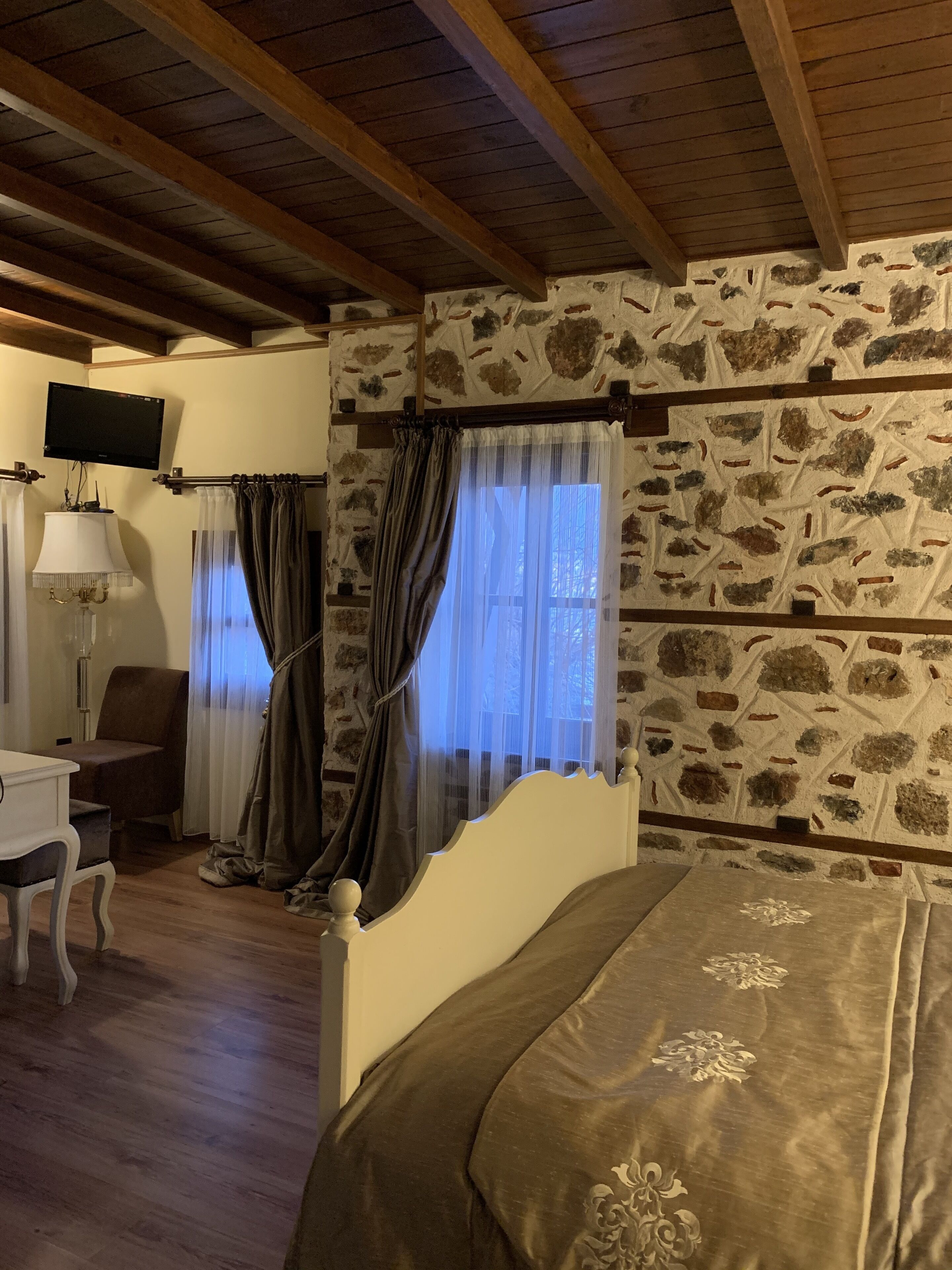 Ein Zimmer mit Steinwand, Holzbalken, einem Bett, einem Schreibtisch, einem Stuhl, einer Lampe und einem Vorhangfenster.