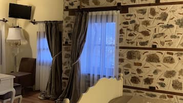 Ein Zimmer mit Steinwand, Holzbalken, einem Bett, einem Schreibtisch, einem Stuhl, einer Lampe und einem Vorhangfenster.