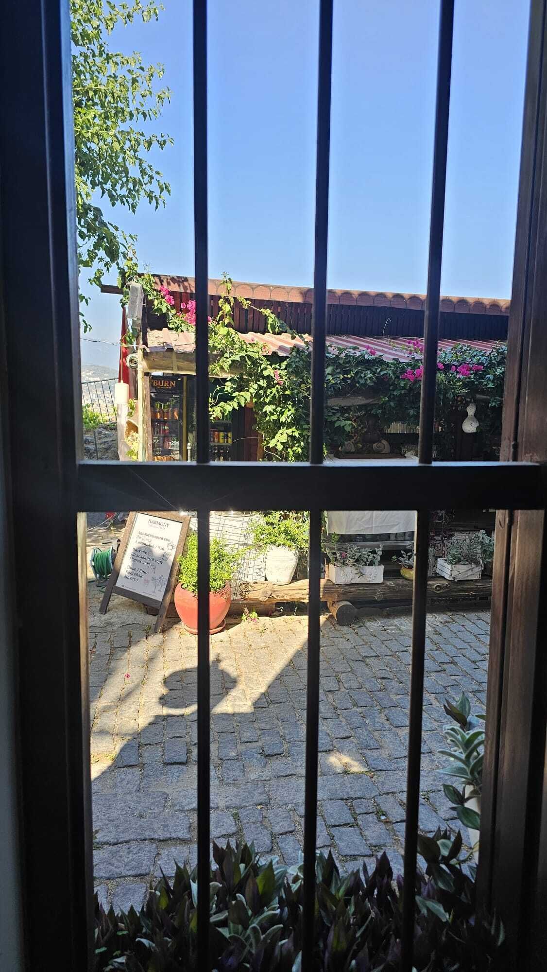 Blick durch ein Fenster mit Metallgitter auf einen Innenhof mit Holzzaun, Blumentöpfen und einem Schild.