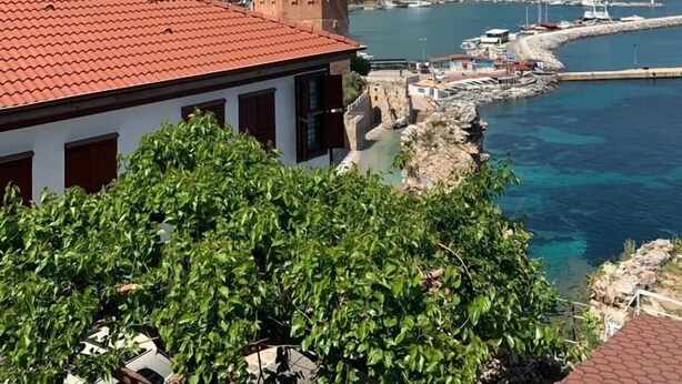 A coastal cityscape with a prominent red-roofed building in the foreground, a marina with boats, and a mountainous backdrop under a clear sky. - Harmony Boutique Hotel (Alanya)