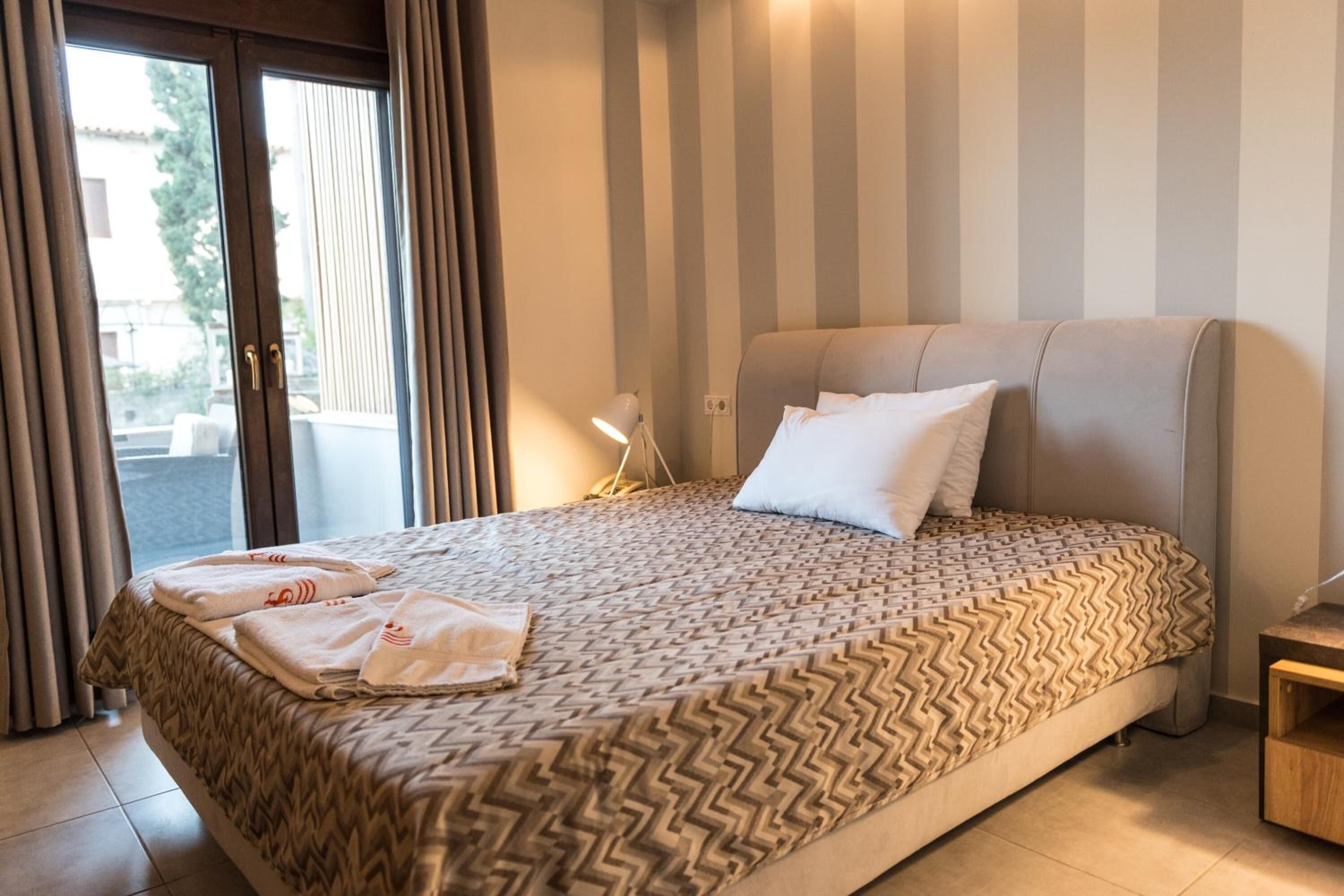 Een hotelkamer met een bed, nachtkastje, lamp en uitzicht door een raam.