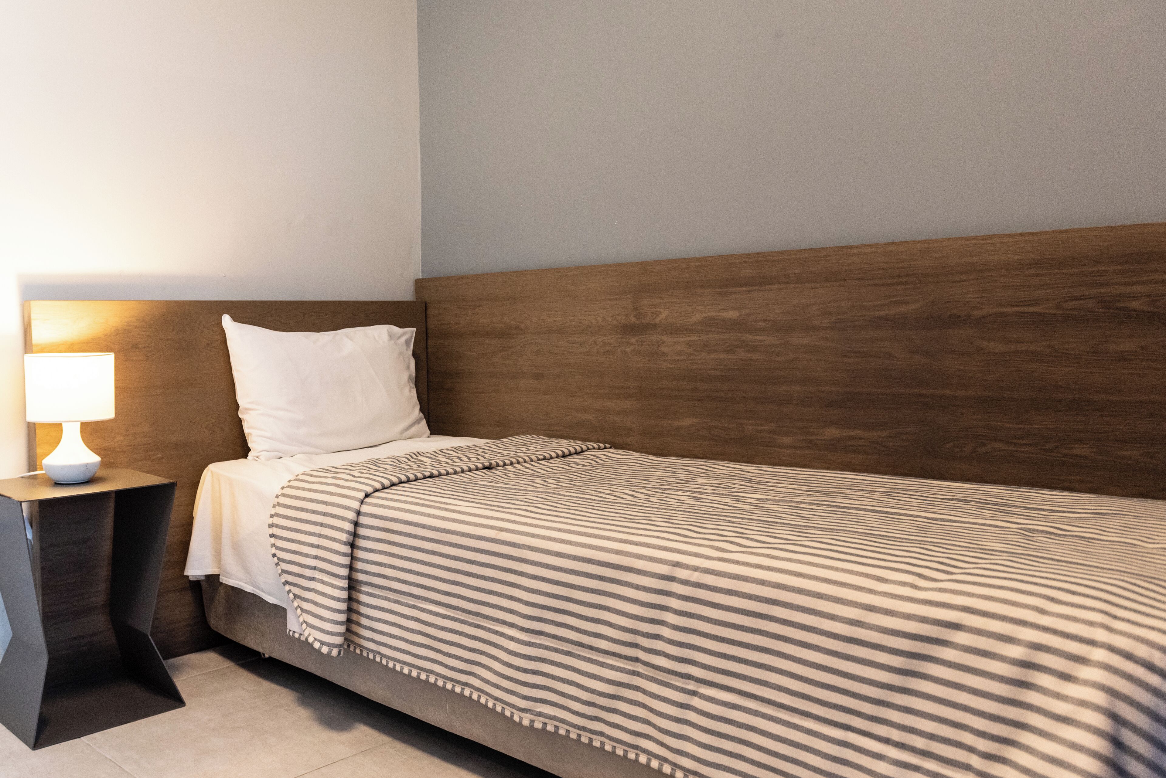 Un lit simple avec une tête de lit en bois, une table de chevet avec une lampe et une couverture à rayures.