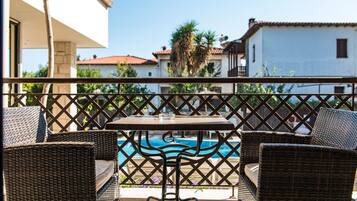 A balcony with wicker furniture, a table, and a view of a swimming pool and buildings.