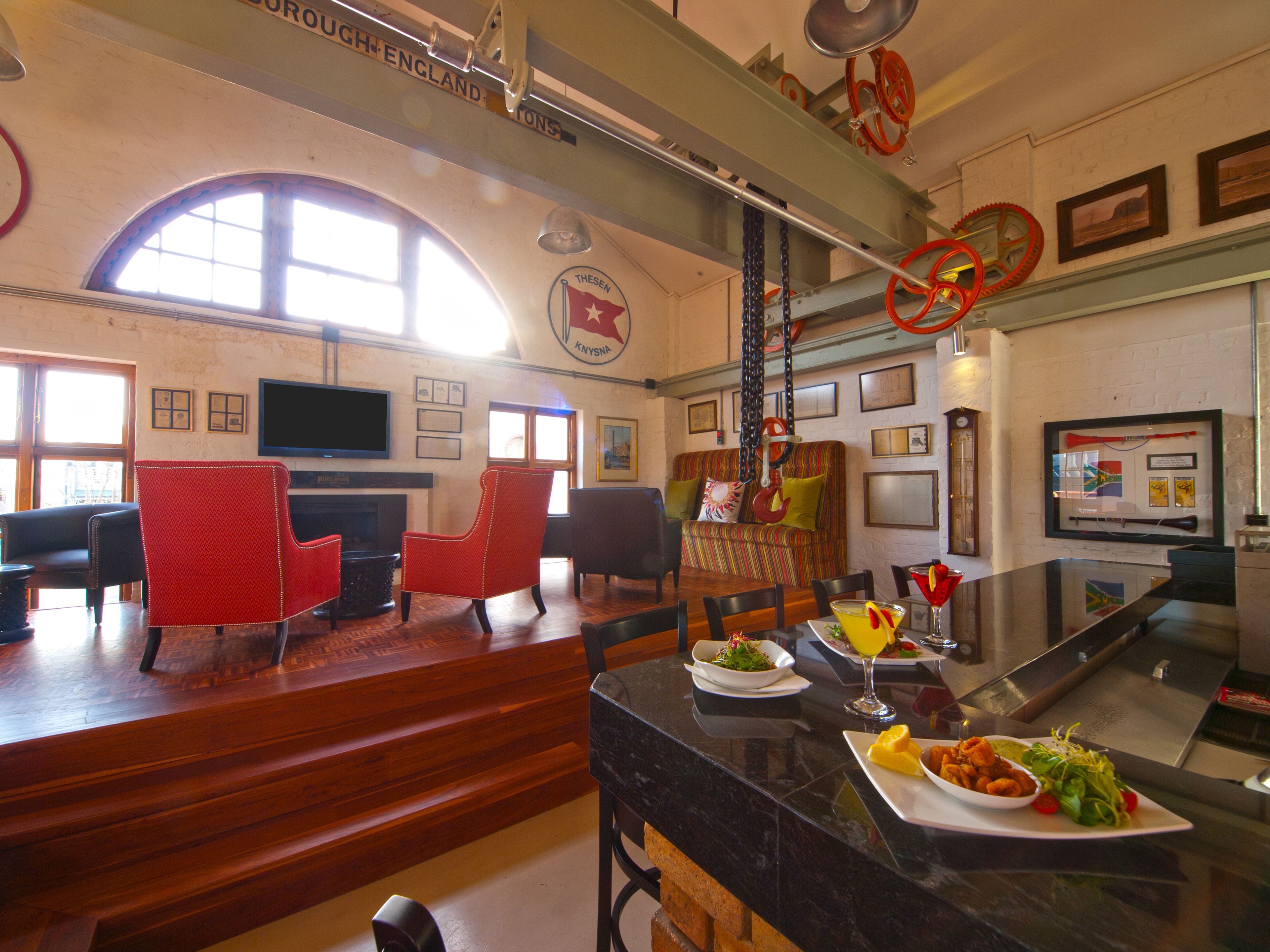 A restaurant with a high ceiling, wooden floors, and red chairs. There is a bar area with a counter and a television mounted on the wall.