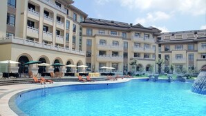 Una gran piscina con sillones de playa y sombrillas, rodeada por un edificio de varios pisos con balcones.