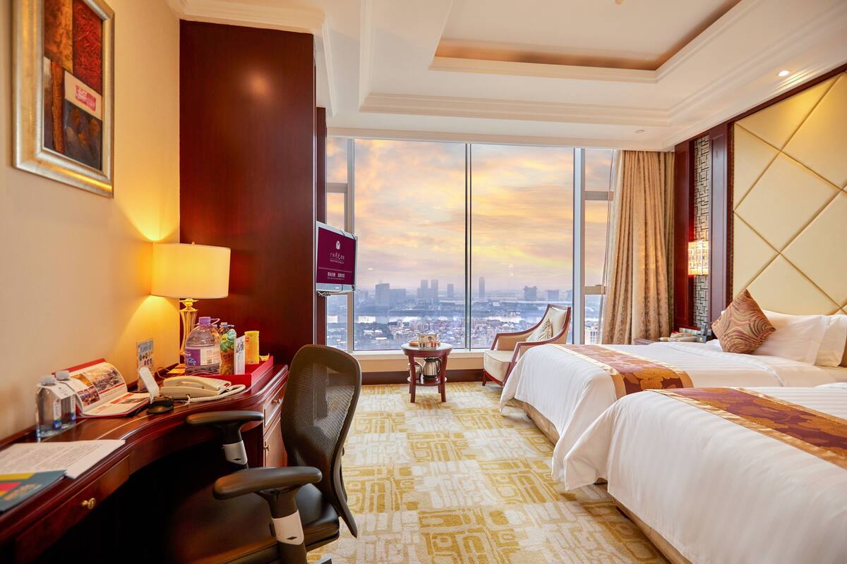 A hotel room with two beds, a desk with a lamp, a chair, and a view of the cityscape.