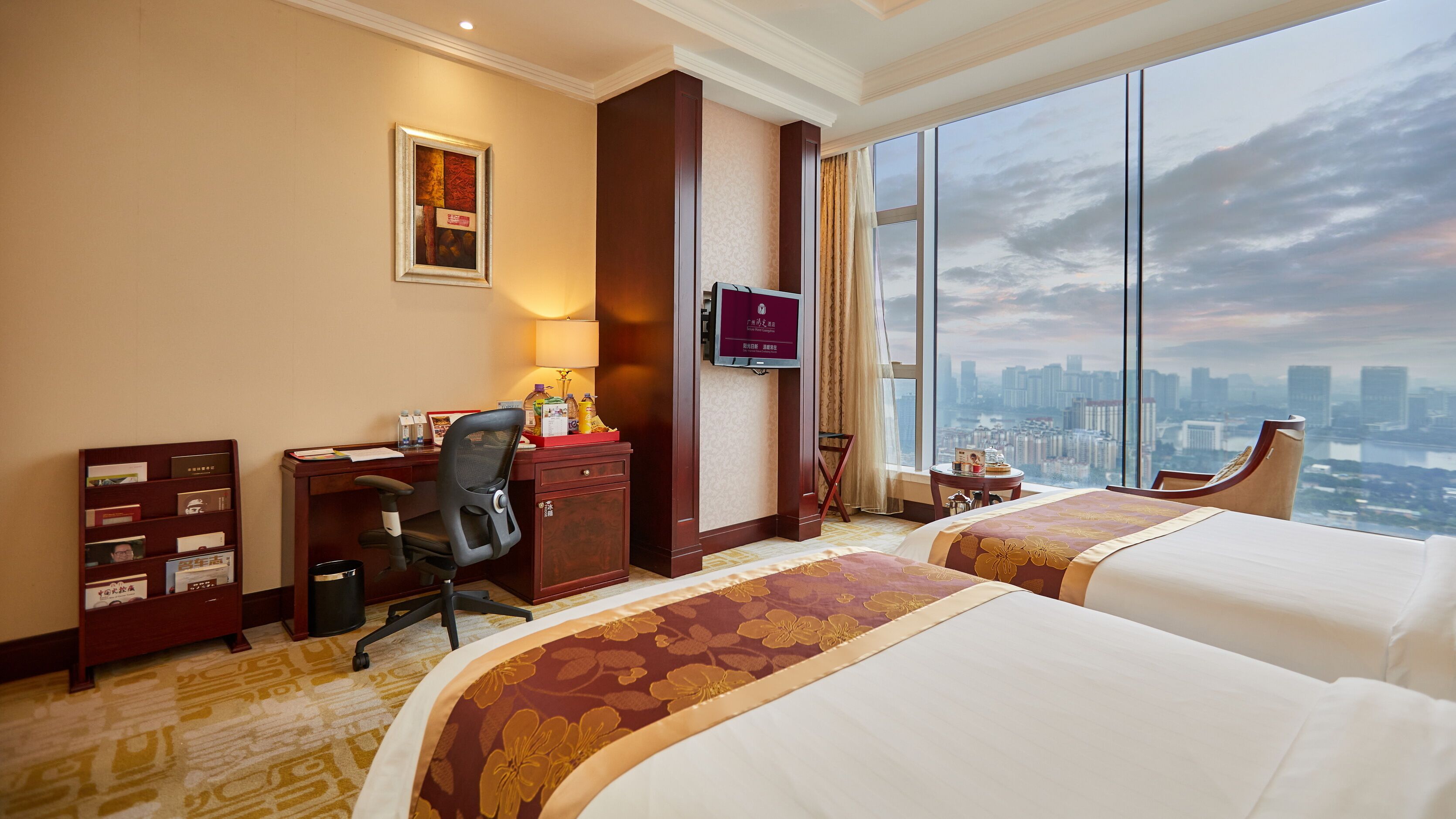 A hotel room with two beds, a desk with a chair, a TV, and a large window with a city view.