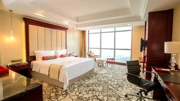 A hotel room with a large bed, a desk, a chair, and a view of the city through large windows.