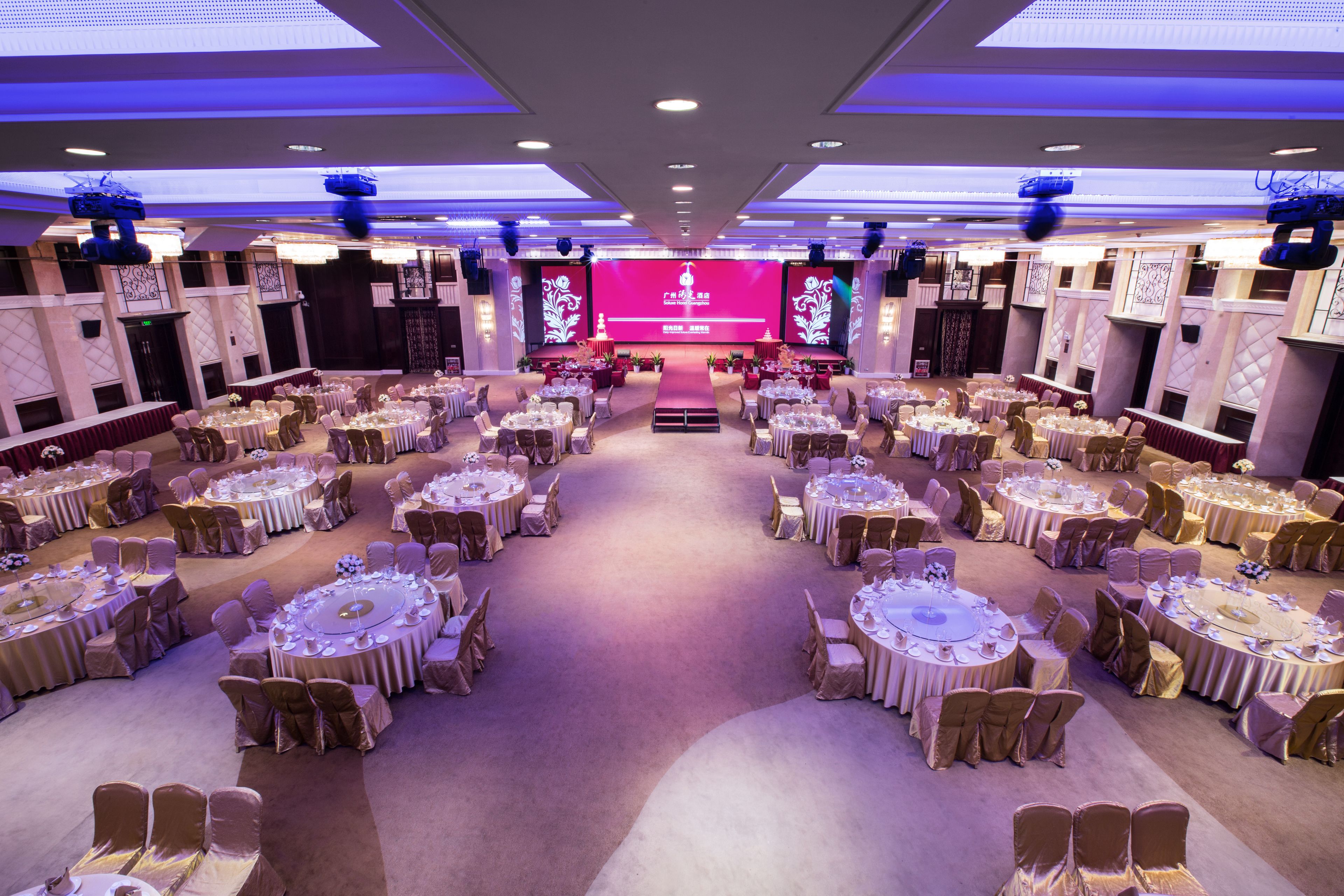 Uma sala de banquetes com mesas e cadeiras redondas, um palco com uma tela grande e piso coberto de carpete.
