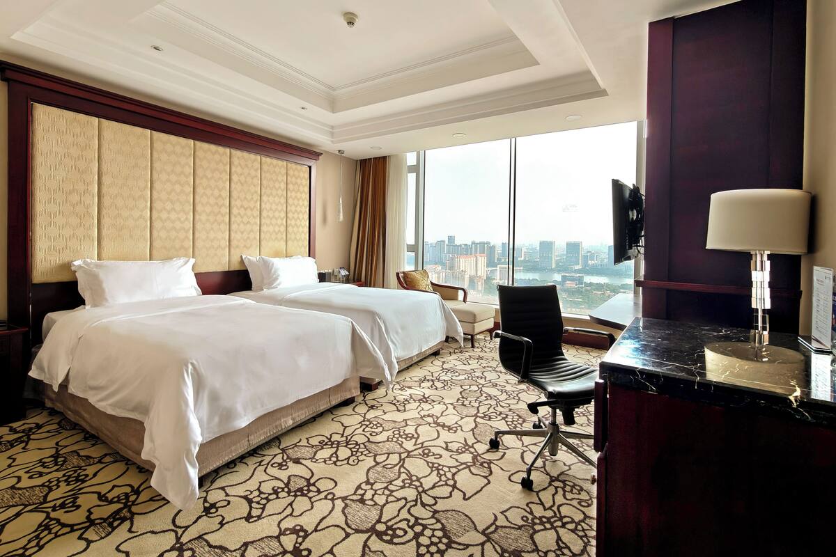A hotel room with two beds, a desk, a chair, a TV, and a large window with a city view.