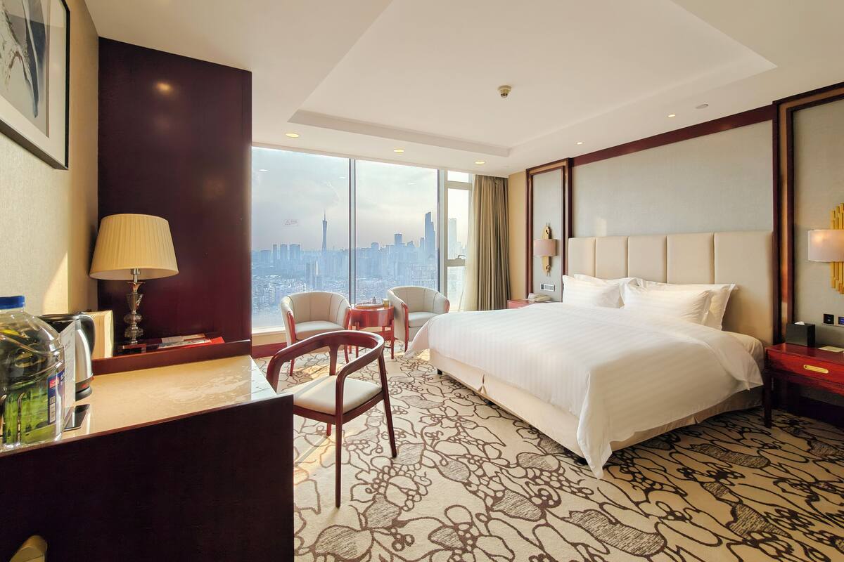 A hotel room with a large bed, a desk with a lamp, a chair, and a view of a cityscape through large windows.