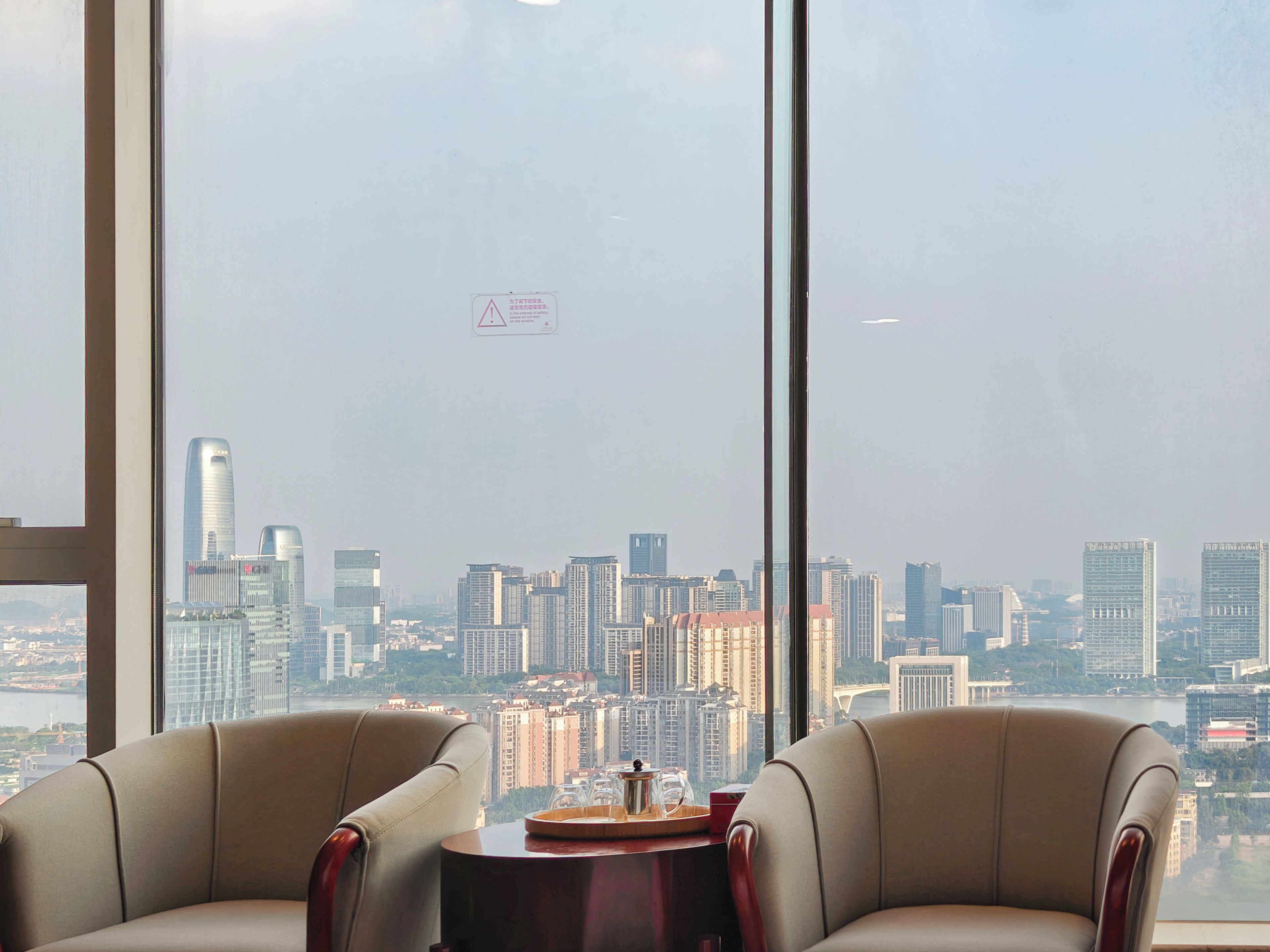 A room with two leather chairs, a small round table, and a view of a cityscape through large windows.