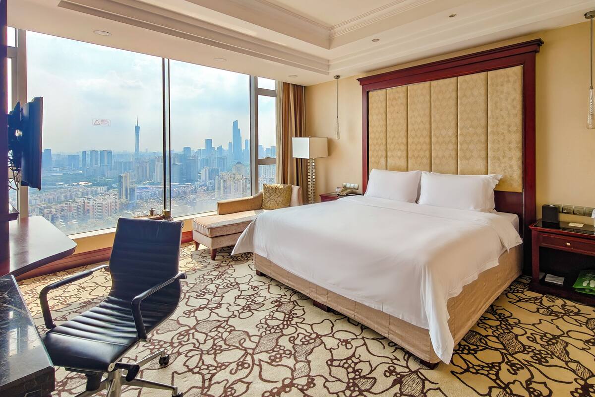 A hotel room with a large bed, a desk, a chair, a nightstand, and a view of the cityscape.