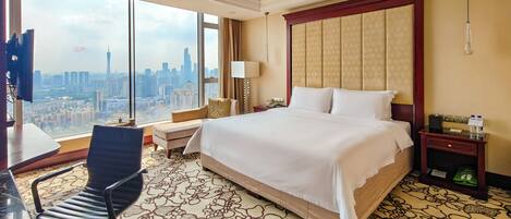 A hotel room with a large bed, a desk, a chair, a nightstand, and a view of the cityscape.