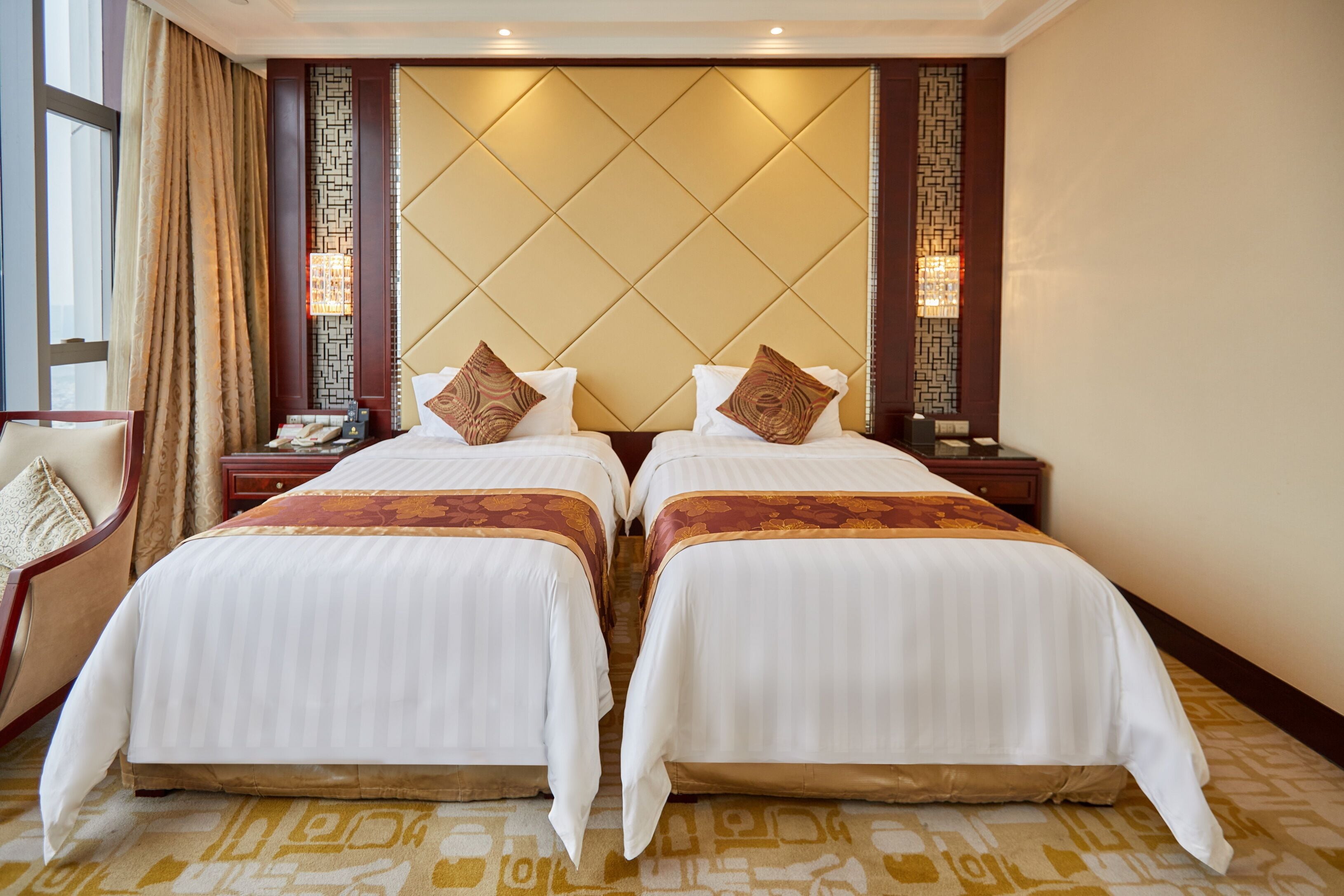 A hotel room with two beds, a large window with curtains, a nightstand with a phone, and a wall with a geometric pattern.
