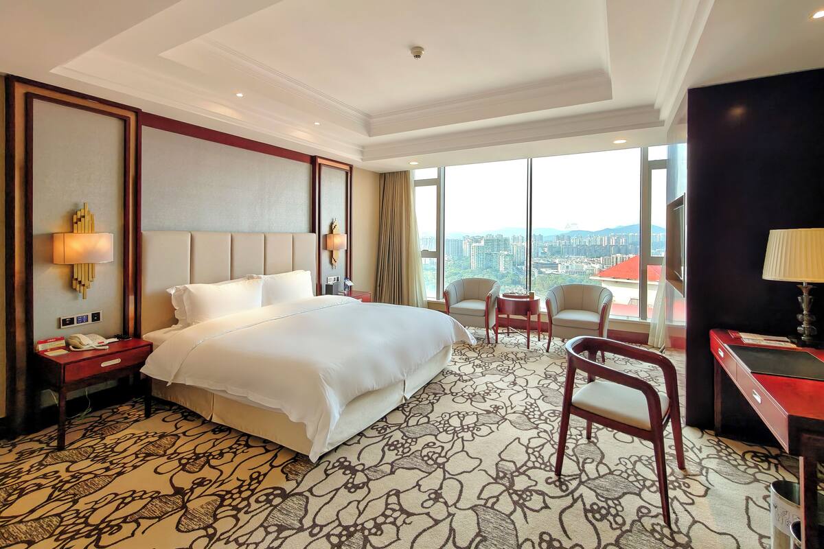A hotel room with a large bed, a desk, two chairs, a large window with a city view, and a patterned carpet.