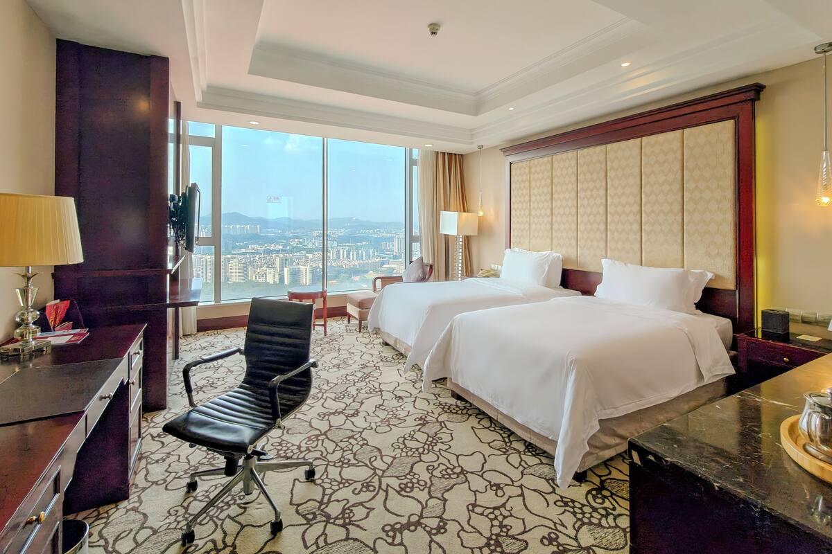 A hotel room with two beds, a desk, a chair, and a large window offering a city view.