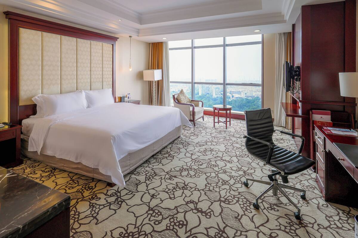A hotel room with a large bed, a desk, a chair, and a view of the city through large windows.