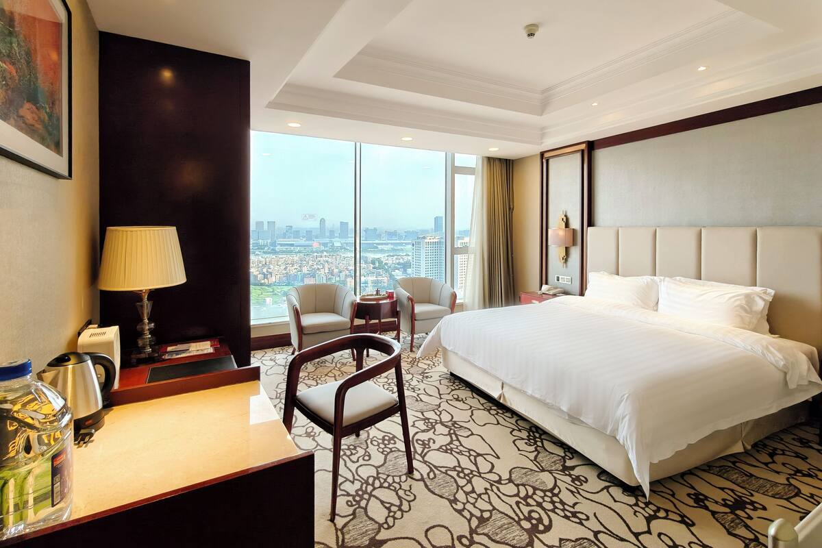 A hotel room with a large bed, two armchairs, a desk with a lamp, and a view of the city.