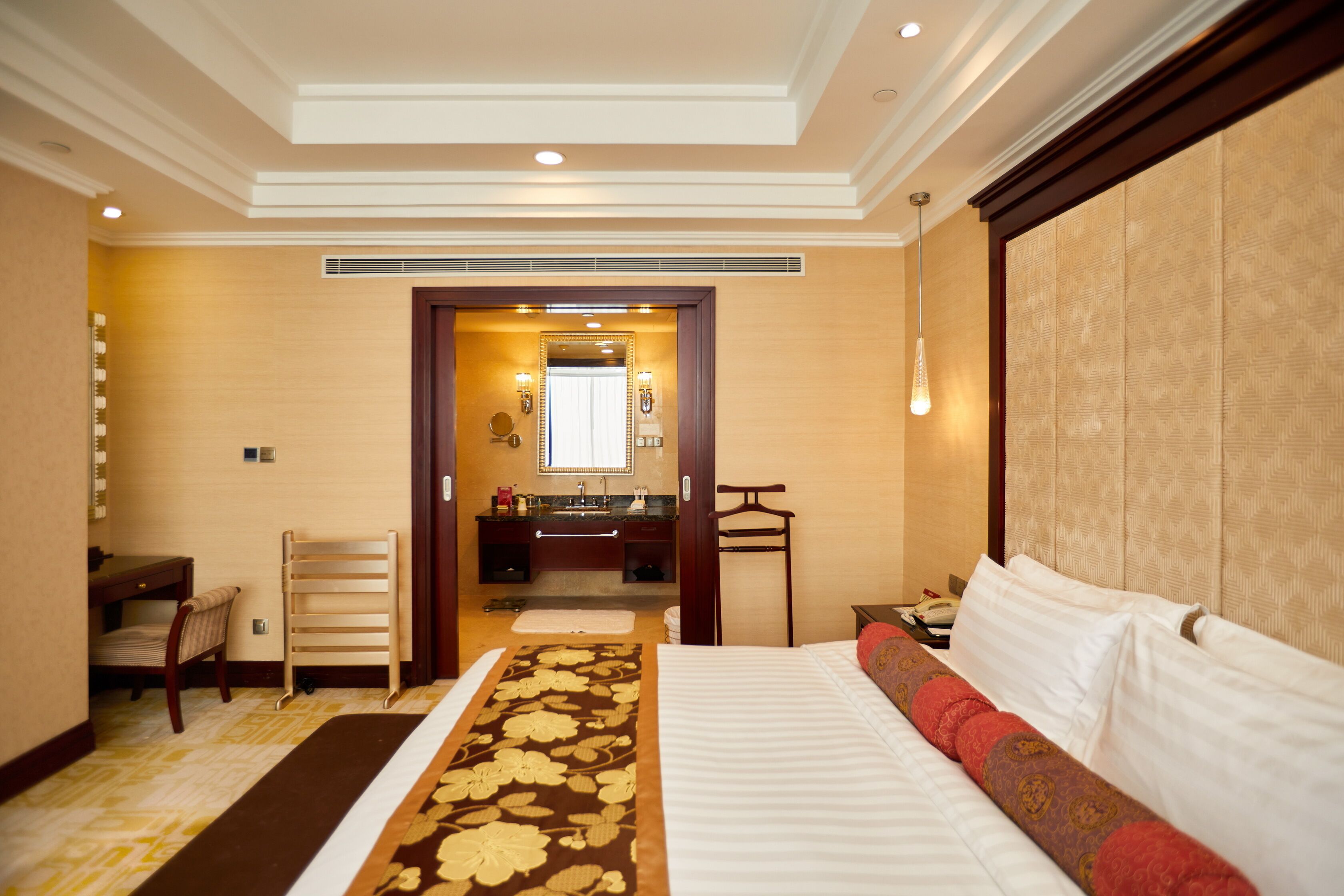 A hotel room with a large bed, a desk, a chair, a vanity with a mirror, and a hanging light fixture.