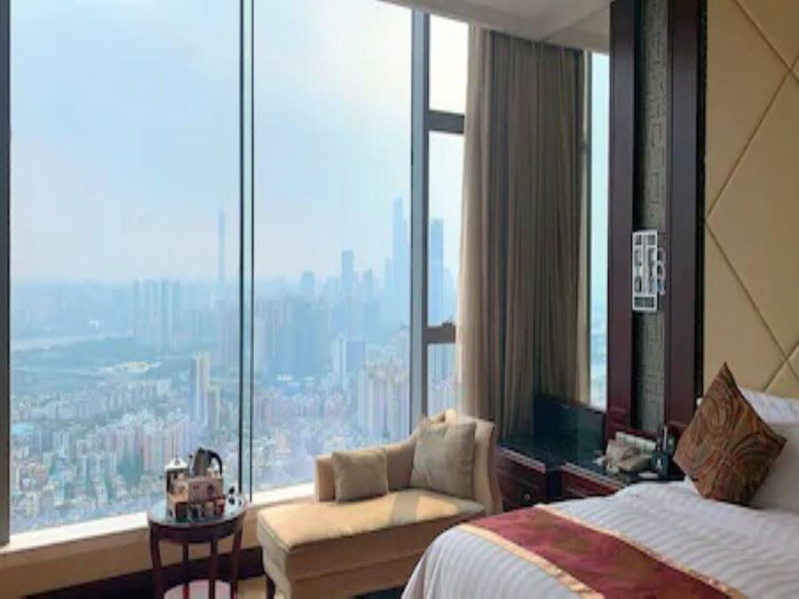 Habitación de hotel con un ventanal amplio que ofrece vistas a la ciudad, una cama, un sofá y una mesita de noche con una lámpara.
