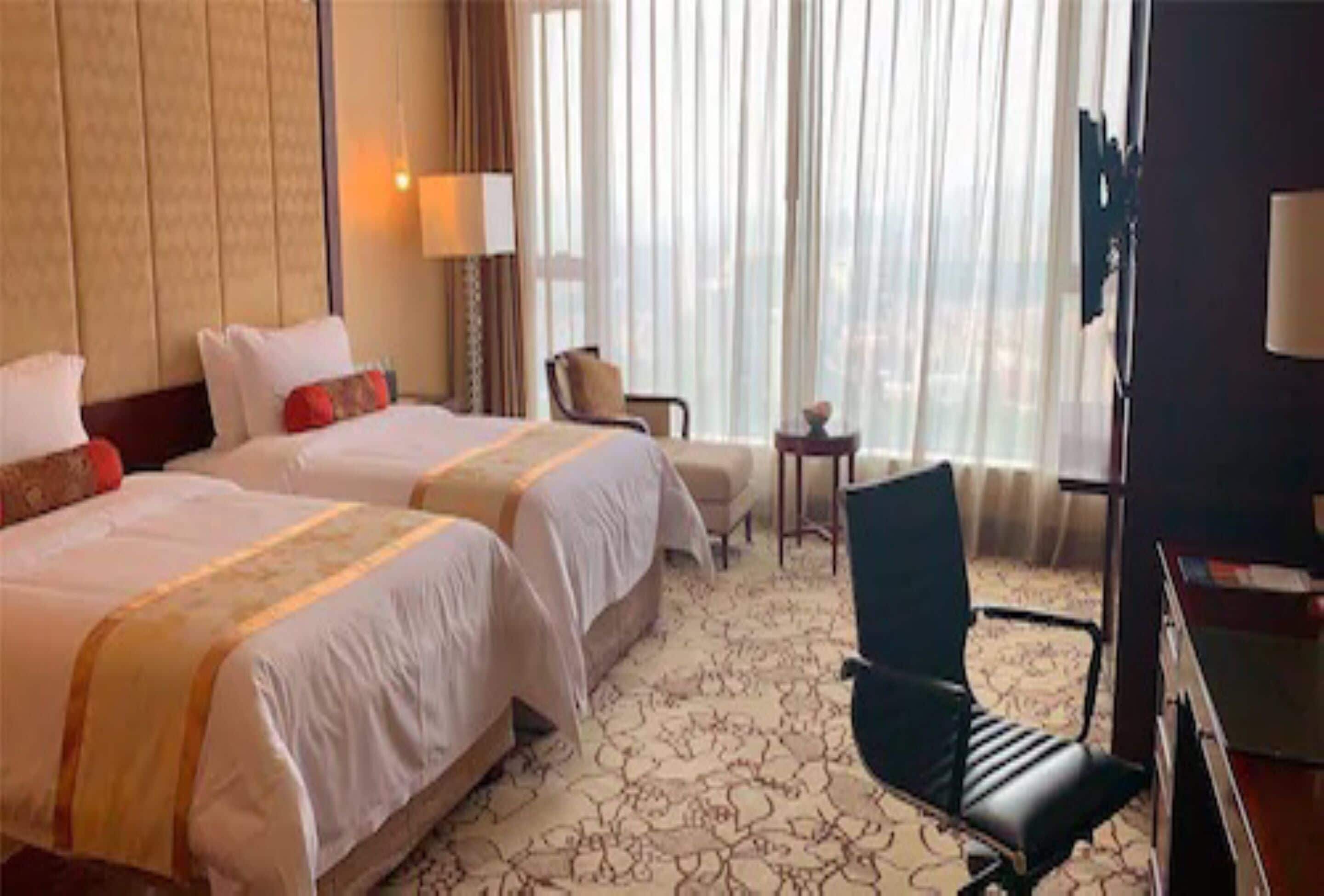 Hotelkamer met twee bedden, een bureau, een stoel en uitzicht op de stad door grote ramen.