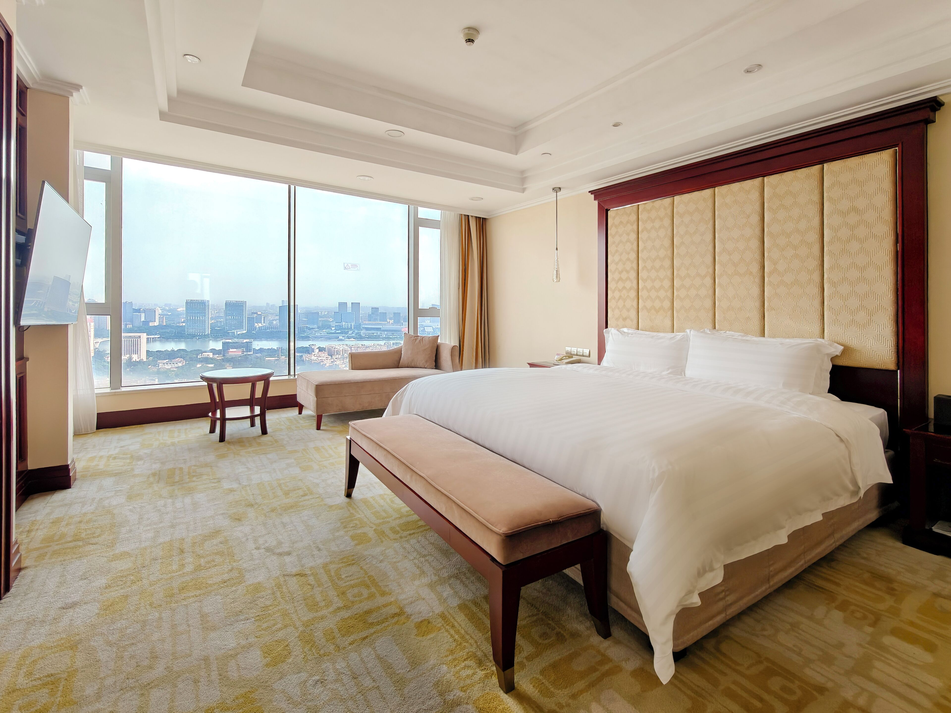 A hotel room with a large bed, a bench, a small table, and a view of a cityscape through large windows.