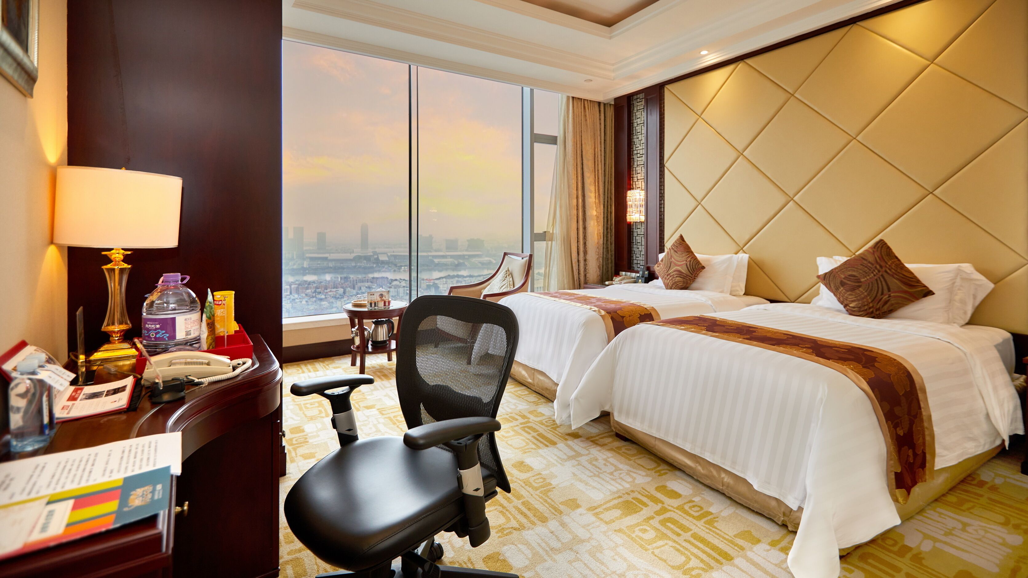Une chambre d’hôtel avec deux lits, un bureau avec une lampe, une chaise et une vue sur le paysage urbain grâce à de grandes fenêtres.