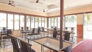 A spacious indoor area with multiple tables and chairs, large windows, and ceiling fans. - KTDC Nandanam Guruvayoor (Chavakkad)
