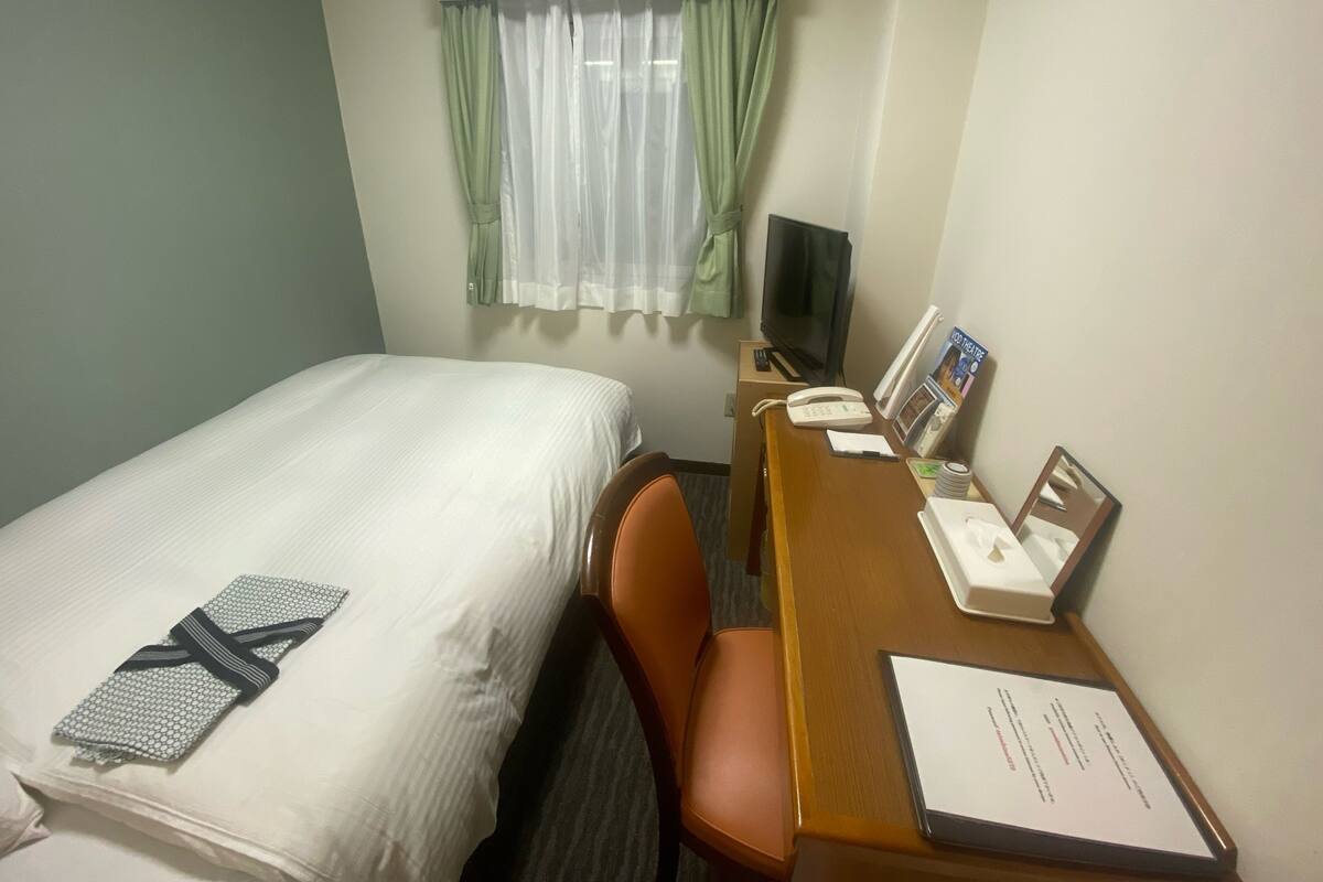 A hotel room with a bed, a desk with a telephone, a television, and a window with curtains.