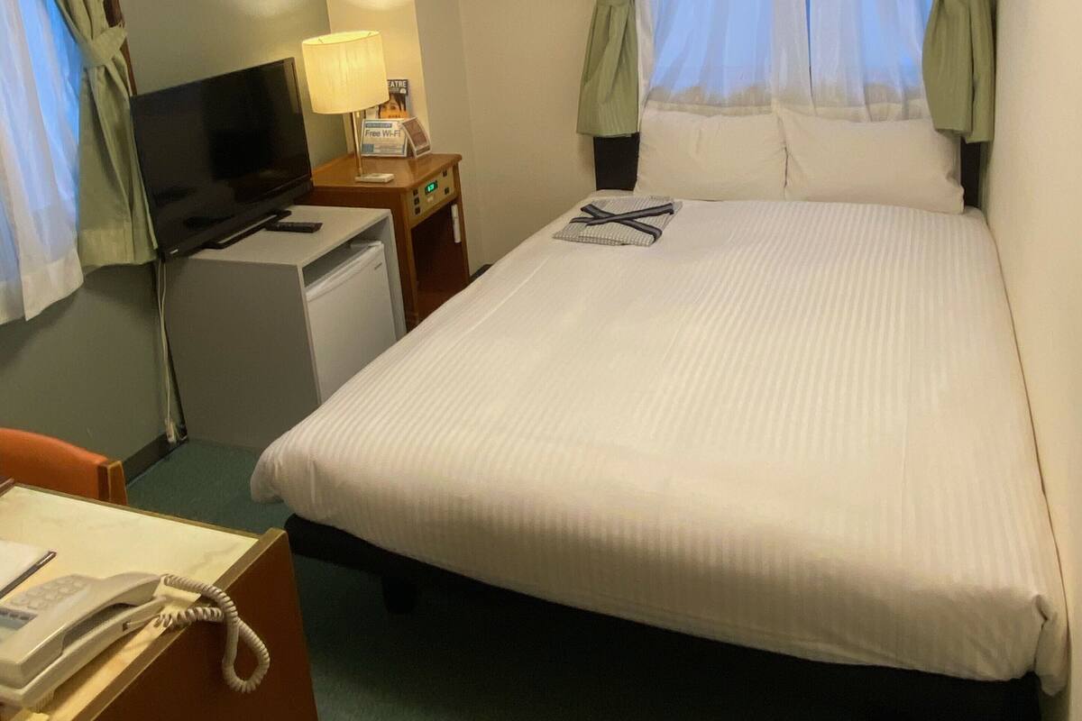 A hotel room with a bed, a desk with a computer, an air conditioning unit, a telephone, and a painting on the wall.