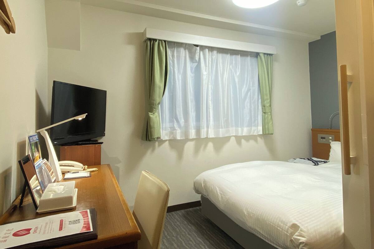 A hotel room with a bed, a desk with a telephone and a television, a window with curtains, and a bedside table with a lamp and a tissue box.