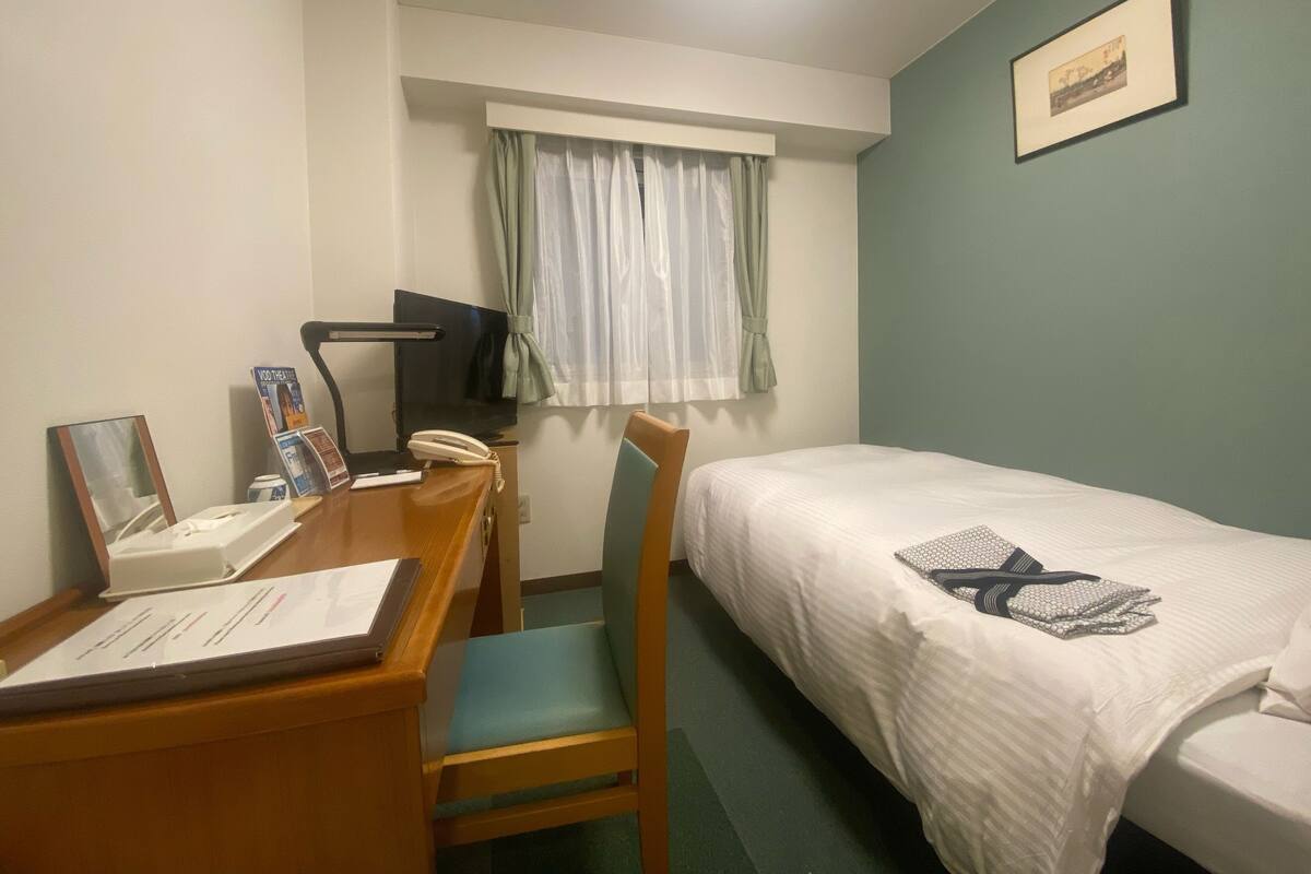 A hotel room with a single bed, a desk with a chair, a television, and a window with curtains.
