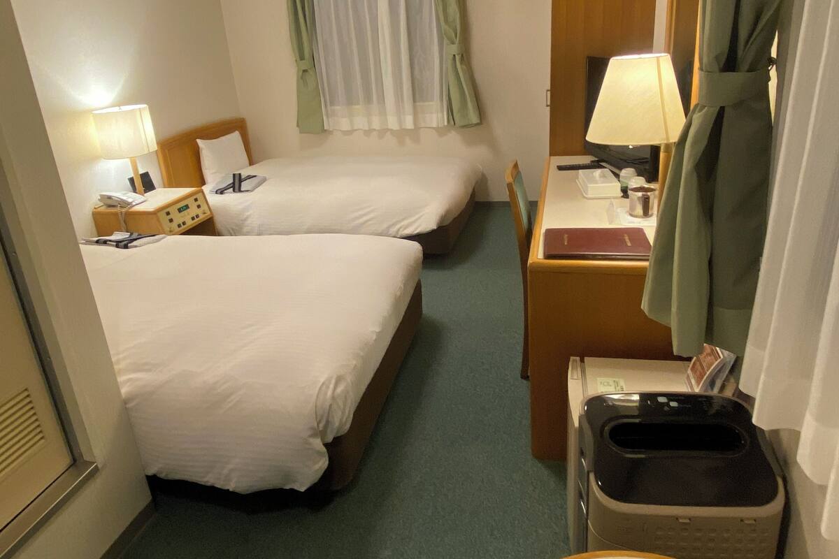 A hotel room with two beds, a desk, a chair, a telephone, a lamp, a robe, and a trash bin.