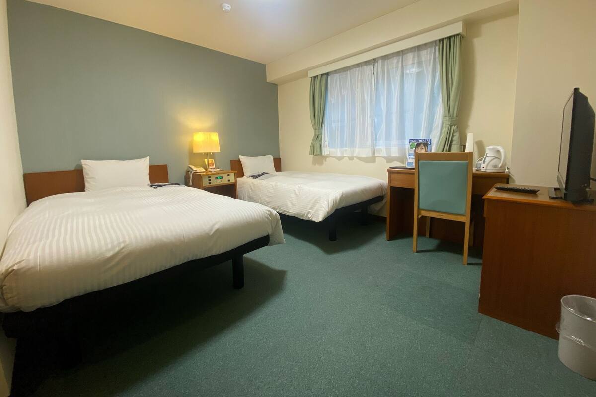 A hotel room with two beds, a desk with a computer, a chair, a TV, and a window with curtains.