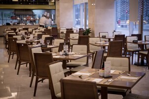 A spacious restaurant with multiple tables and chairs, a visible kitchen area, and large windows allowing natural light.