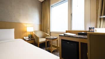 A hotel room with a bed, a chair, a small table, a desk with a computer, a mini-fridge, and a window with curtains.