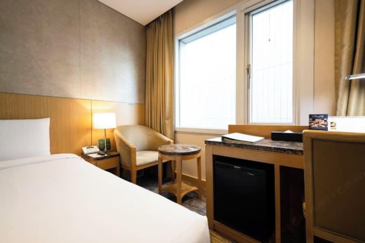 Habitación de hotel con cama, escritorio con computadora, silla, mesita y ventana con cortinas.